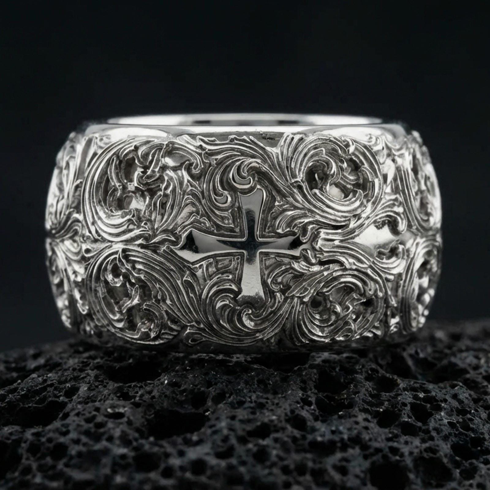 Polished silver ornate cross band ring on volcanic stone, crisp product photo emphasizing the bright reflective finish and detailed baroque engravings.