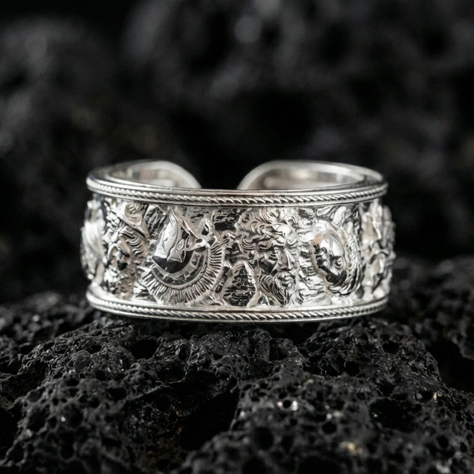 Polished silver mythic faces cuff band ring on volcanic stone, crisp product photo emphasizing clean shine and high-definition sculpted faces.
