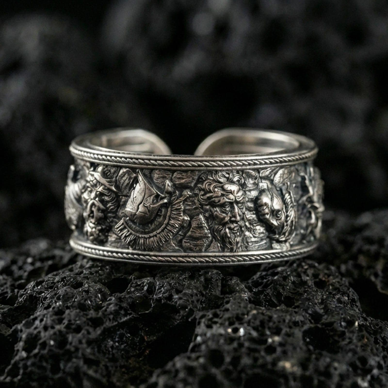 Oxidized sterling silver mythic faces cuff band ring on volcanic stone, macro close-up highlighting deep relief character details and antique contrast.