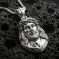 Load image into Gallery viewer, Polished silver Jesus pendant with crown of thorns, flowing hair detail, and raised devotional portrait placed on textured volcanic rock in a premium studio product shot
