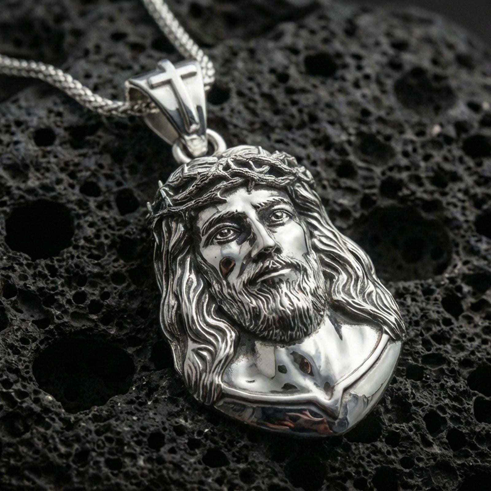 Polished silver Jesus pendant with crown of thorns, flowing hair detail, and raised devotional portrait placed on textured volcanic rock in a premium studio product shot