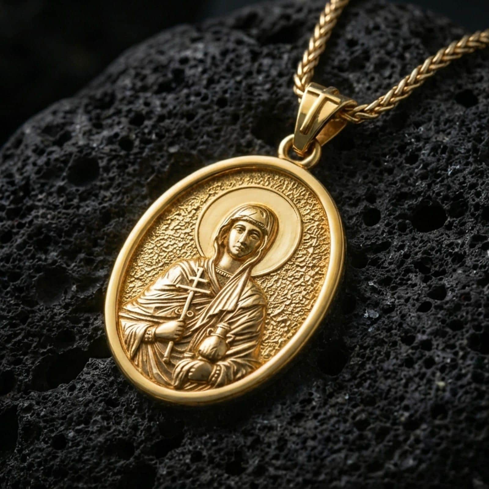 Gold plated Saint Anastasia pendant with detailed saint portrait, sacred cross element, and classic oval medallion design showcased on black volcanic rock in a refined macro jewelry image