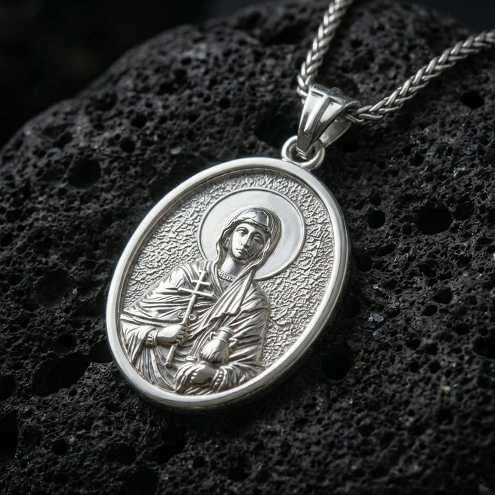 Polished silver Saint Anastasia medallion with engraved orthodox saint relief, halo motif, and oval frame placed on textured volcanic rock in a premium studio product shot