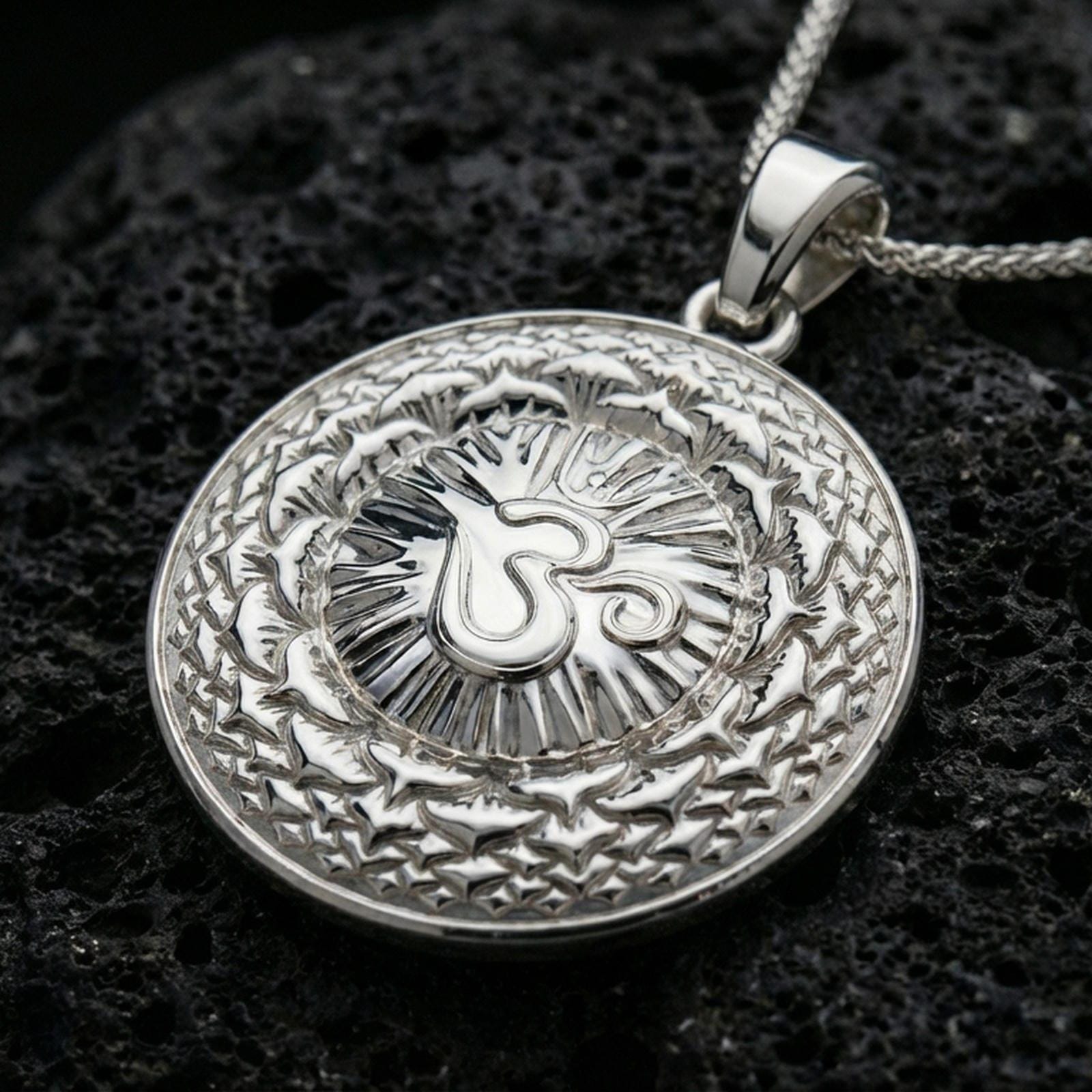 Polished silver spiritual necklace with raised Om symbol, round medallion form, and intricate mandala relief placed on textured volcanic rock in a premium studio product shot