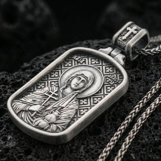 Oxidized Saint Anastasia pendant with haloed saint figure, cross detail, and rounded rectangular orthodox icon design displayed on dark volcanic rock in a close-up luxury jewelry composition