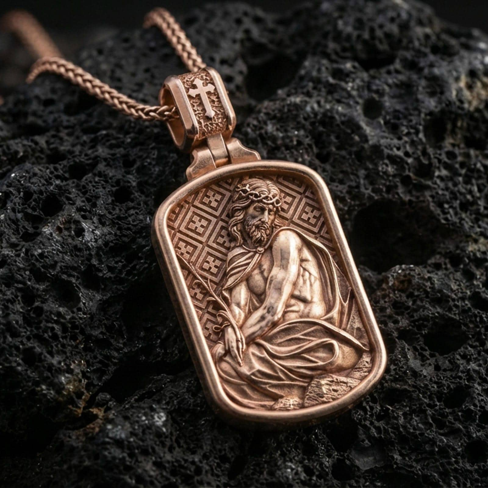 Rose gold plated religious Jesus pendant with seated figure relief, crown of thorns, and rounded rectangular medallion silhouette resting on volcanic rock in an elegant close-up product presentation