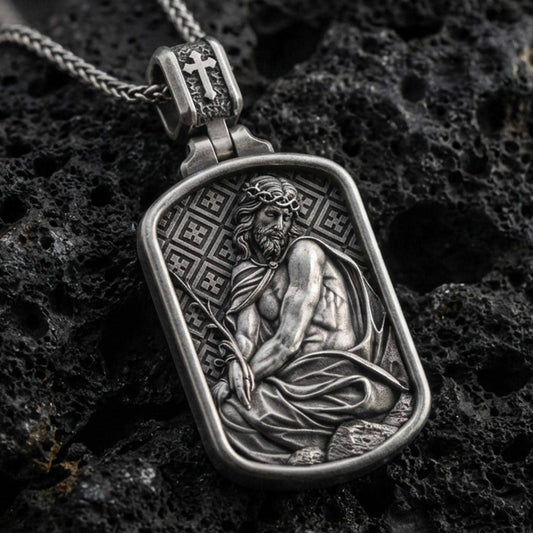 Oxidized Jesus Christ pendant with seated sacred figure, crown of thorns, and framed devotional relief displayed on dark volcanic rock in a close-up luxury jewelry composition