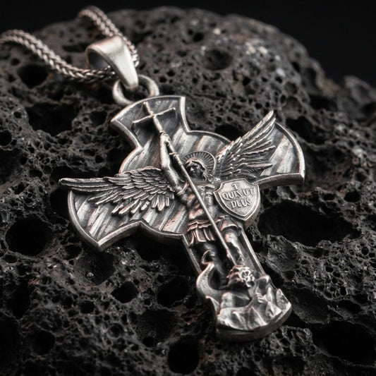 Oxidized Saint Michael pendant with winged archangel figure, Quis Ut Deus shield, and devil-slaying cross design displayed on dark volcanic rock in a close-up luxury jewelry composition