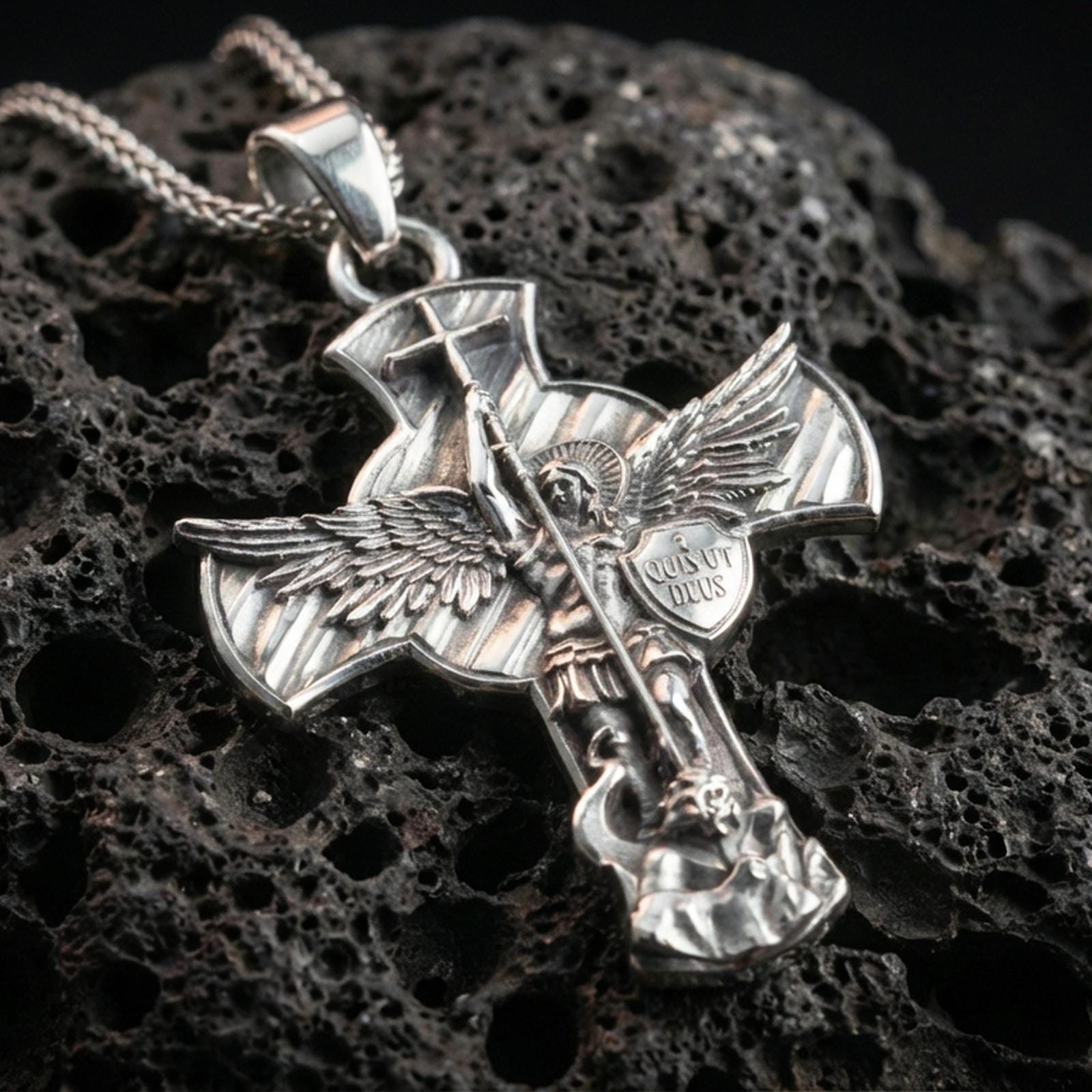 Polished silver Archangel Michael cross pendant with raised warrior relief, shield detail, and dramatic winged silhouette placed on textured volcanic rock in a premium studio product shot
