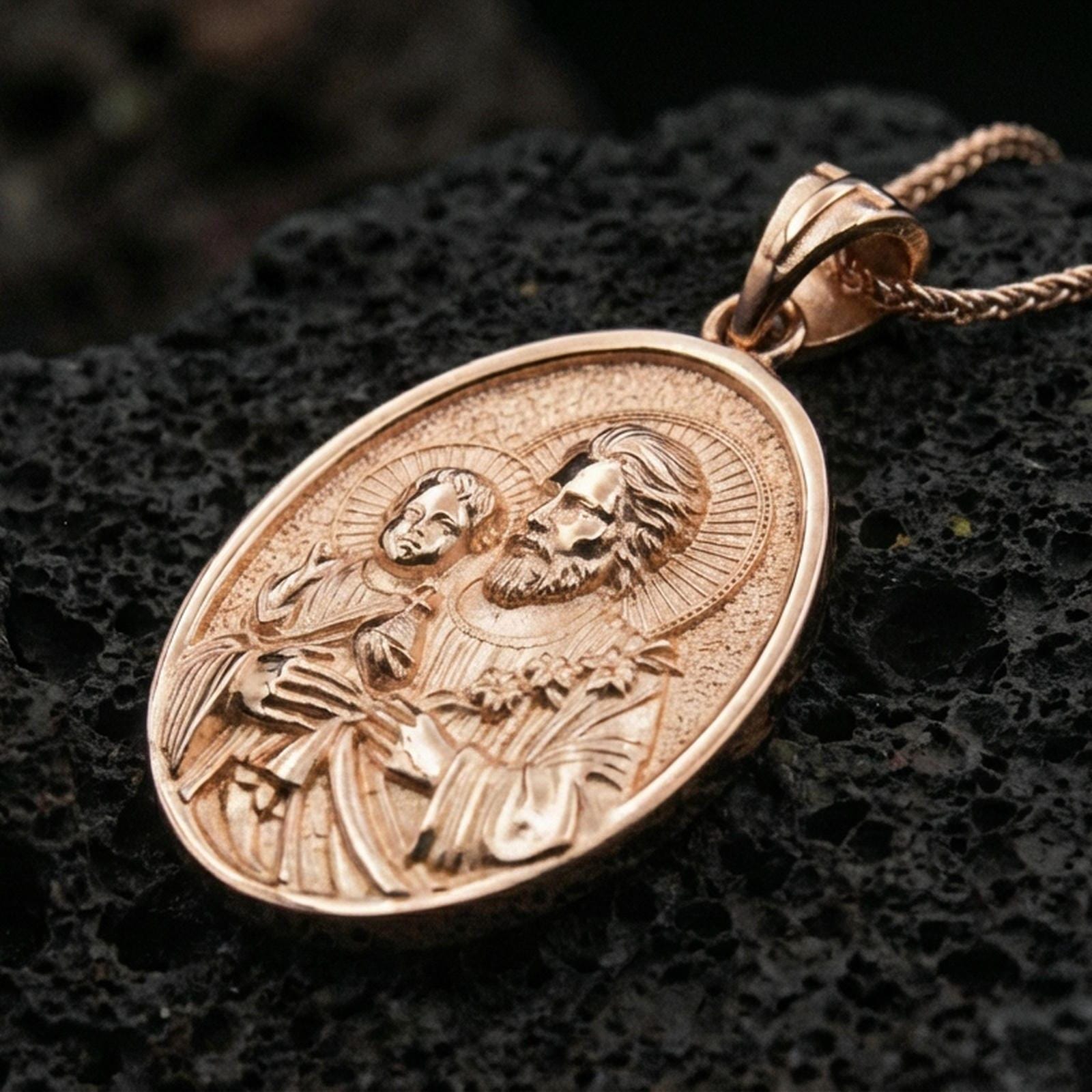 Rose gold plated Saint Joseph necklace with haloed saint portrait, Child Jesus motif, and elegant oval devotional silhouette resting on volcanic rock in an elegant close-up product presentation