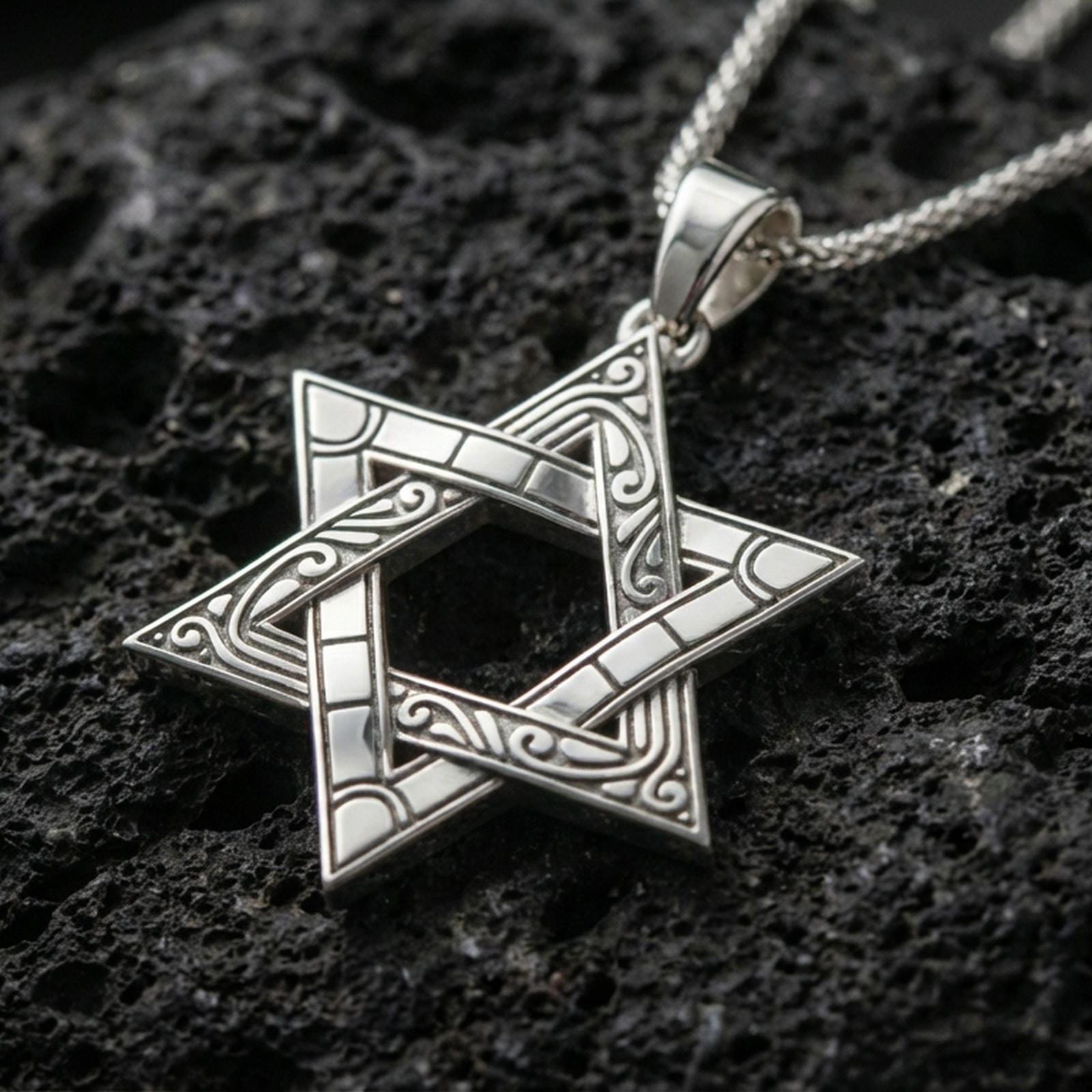 Polished silver Star of David necklace with interlocking six-pointed star design, decorative engraved details, and refined symbolic structure placed on textured volcanic rock in a premium studio product shot