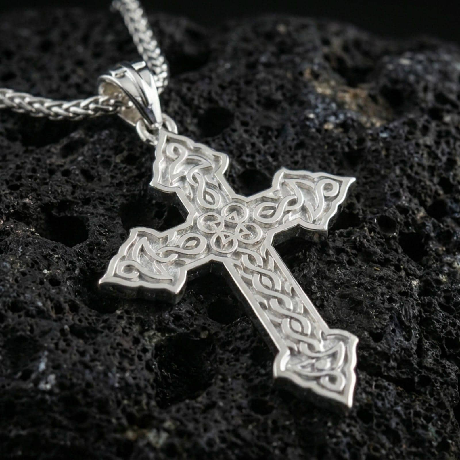 Polished silver cross necklace with detailed interwoven Celtic pattern, pointed cross ends, and refined openwork design placed on textured volcanic rock in a premium studio product shot