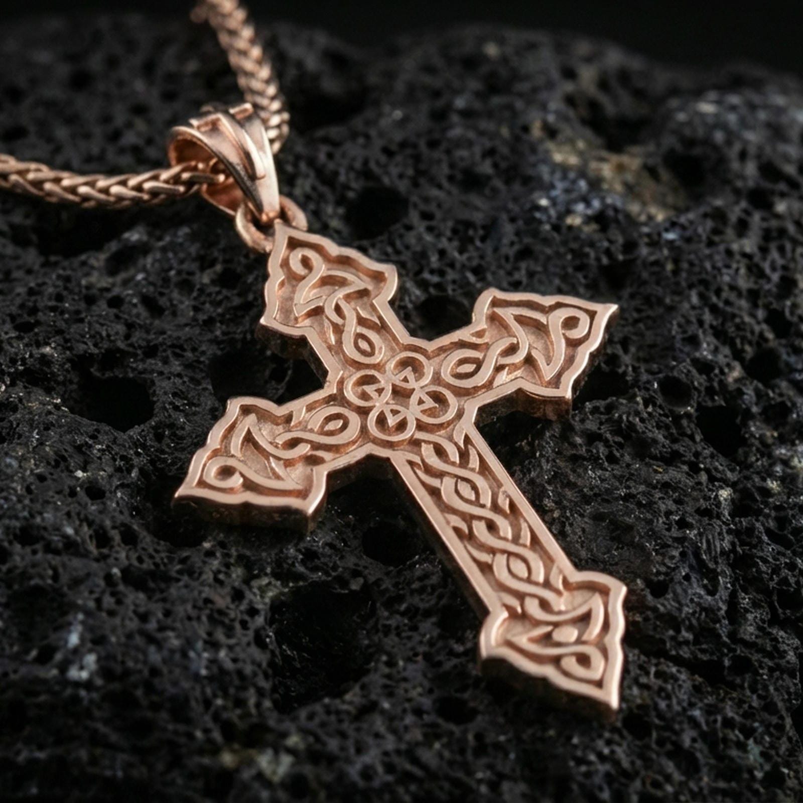 Rose gold plated religious cross pendant with intricate Celtic linework, open center ornament, and elegant pointed silhouette resting on volcanic rock in an elegant close-up product presentation