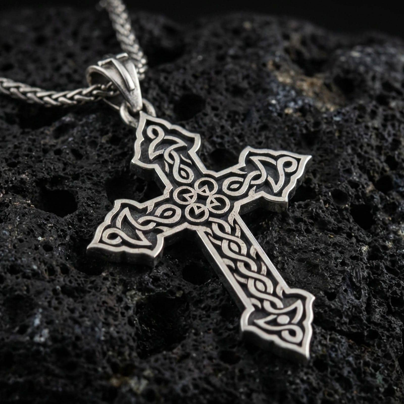 Oxidized Celtic cross pendant with intricate knotwork details and open ornamental silhouette displayed on dark volcanic rock in a close-up luxury jewelry composition