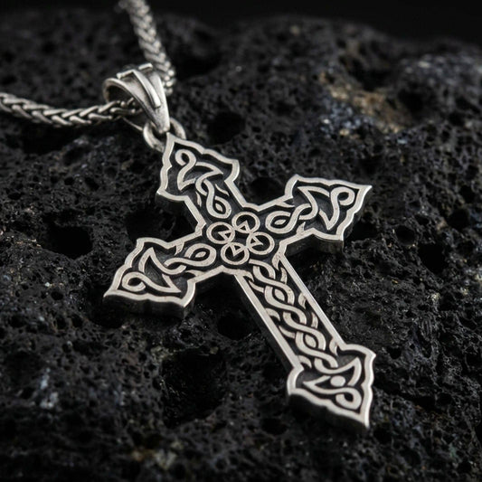 Oxidized Celtic cross pendant with intricate knotwork details and open ornamental silhouette displayed on dark volcanic rock in a close-up luxury jewelry composition