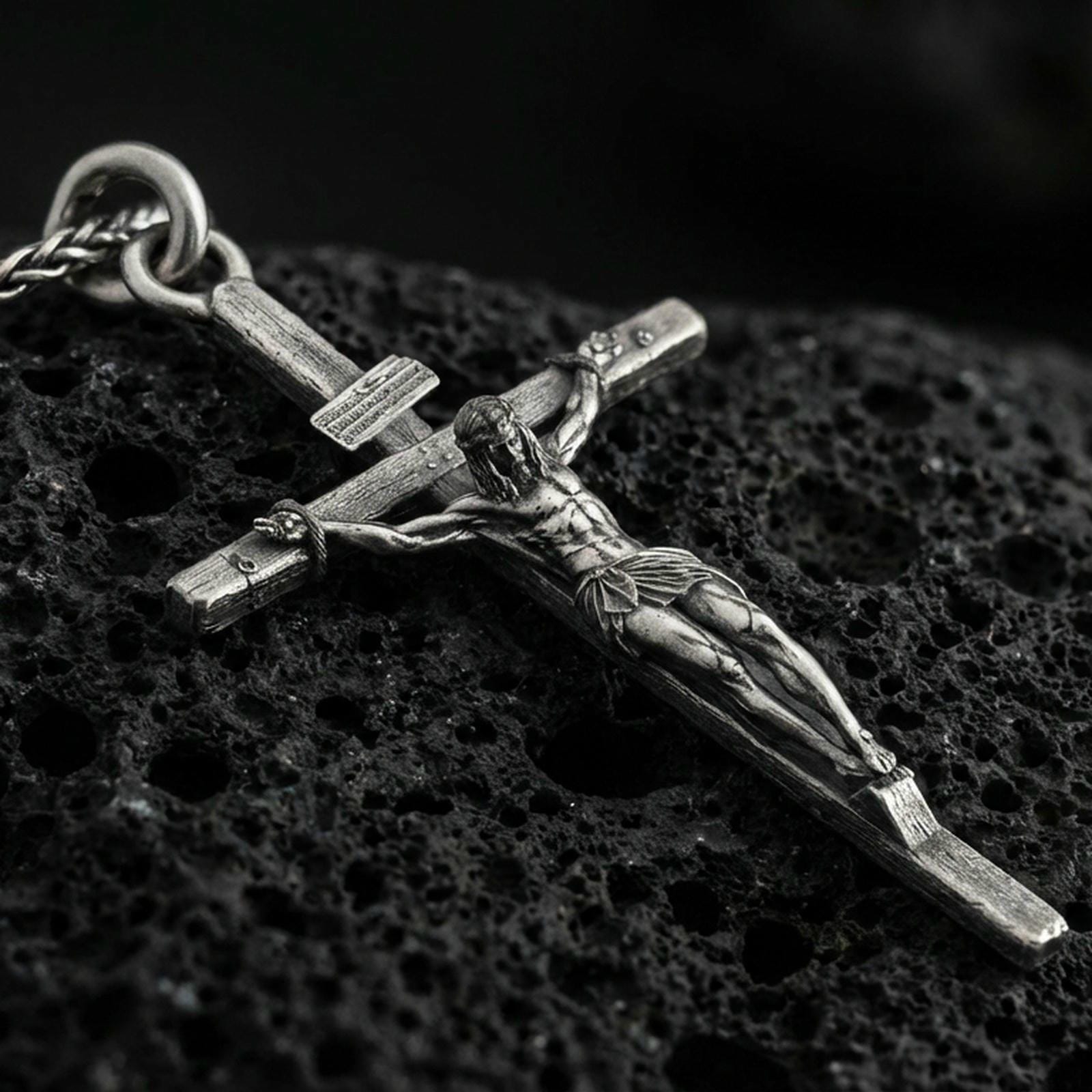 Oxidized sterling silver crucifix pendant on volcanic stone, macro close-up highlighting the deepened relief of the corpus, wood-grain cross texture, and antique contrast.