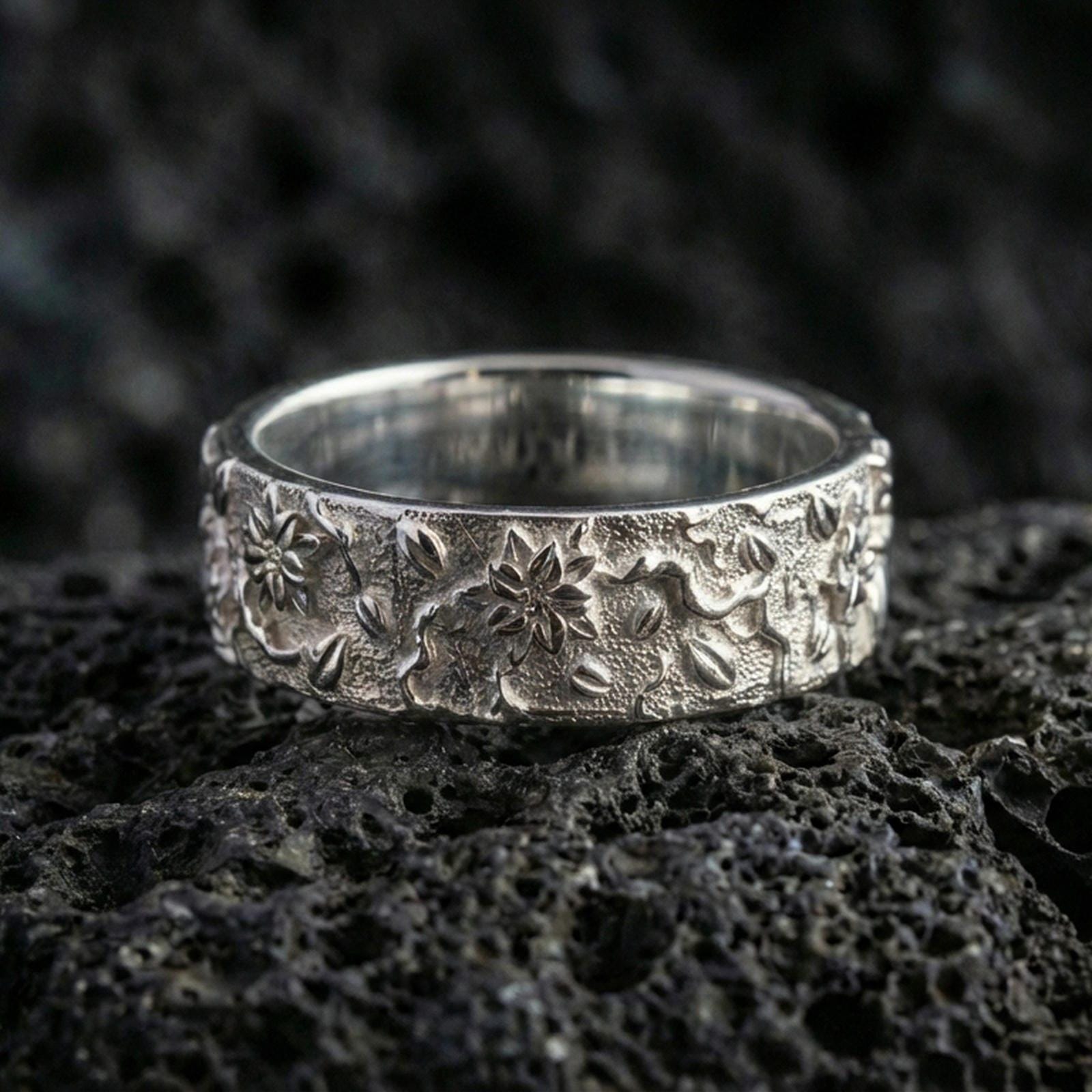 Polished silver floral engraved band ring on volcanic stone, macro shot emphasizing crisp flower relief, smooth shine, and clean botanical detailing.