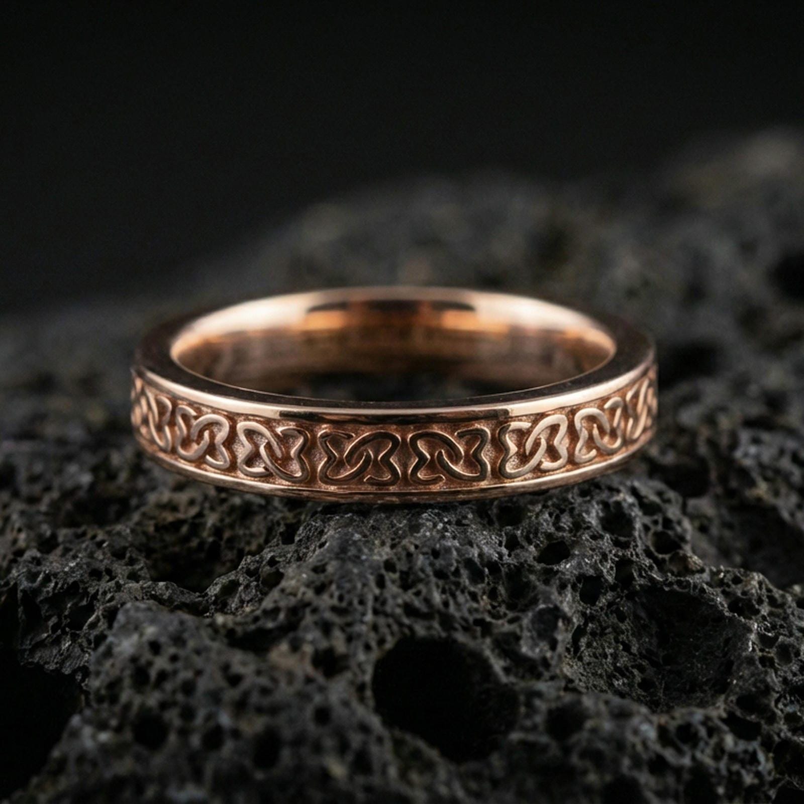 Rose gold plated Celtic knot band ring on volcanic stone, elegant macro photo capturing soft rose reflections and the refined braided motif.