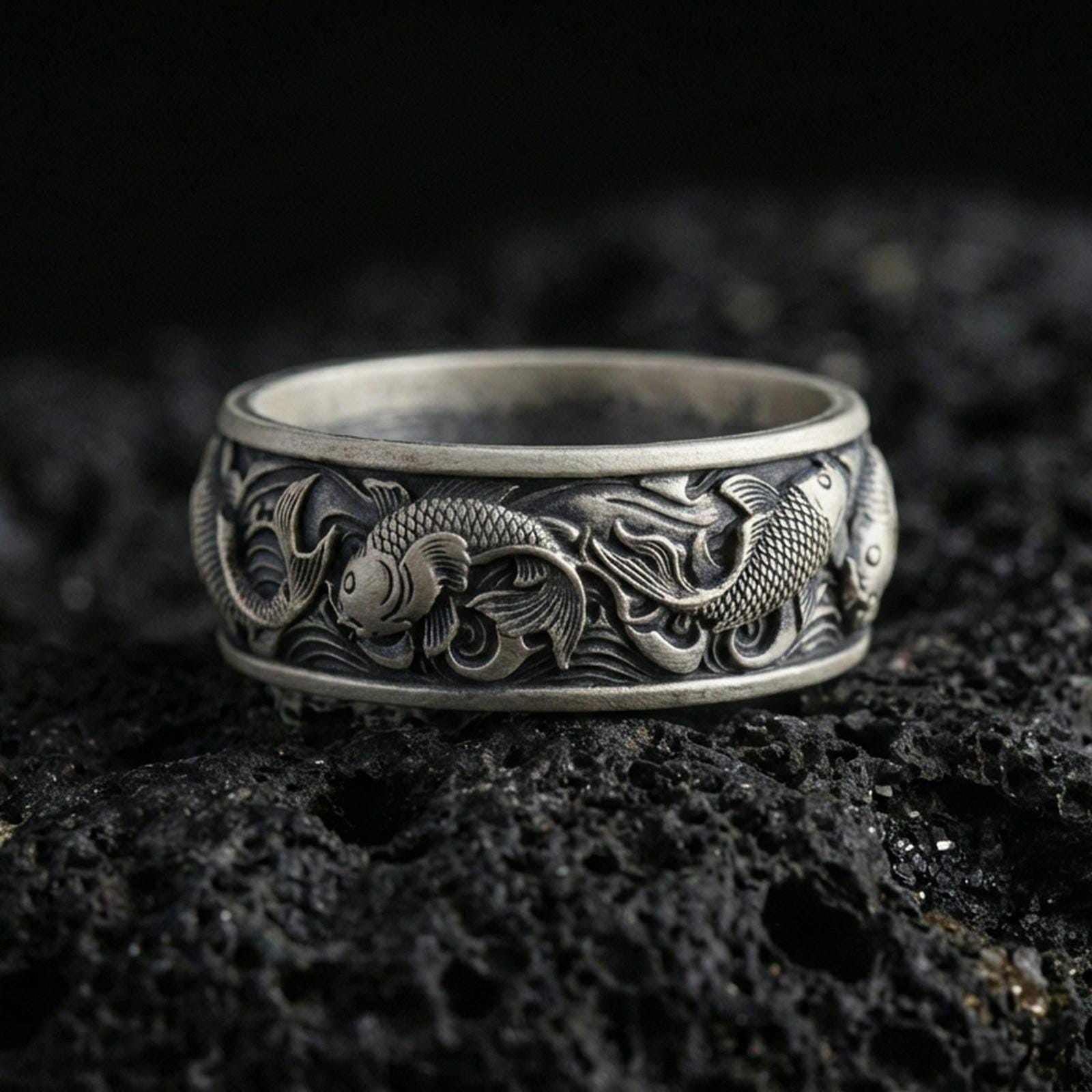 Oxidized sterling silver koi fish band ring on volcanic stone, macro close-up emphasizing the raised koi relief, wave engravings, and antique contrast.