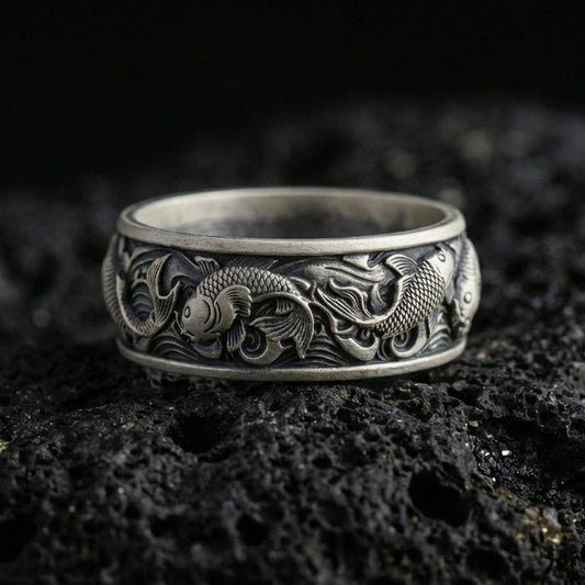 Oxidized sterling silver koi fish band ring on volcanic stone, macro close-up emphasizing the raised koi relief, wave engravings, and antique contrast.