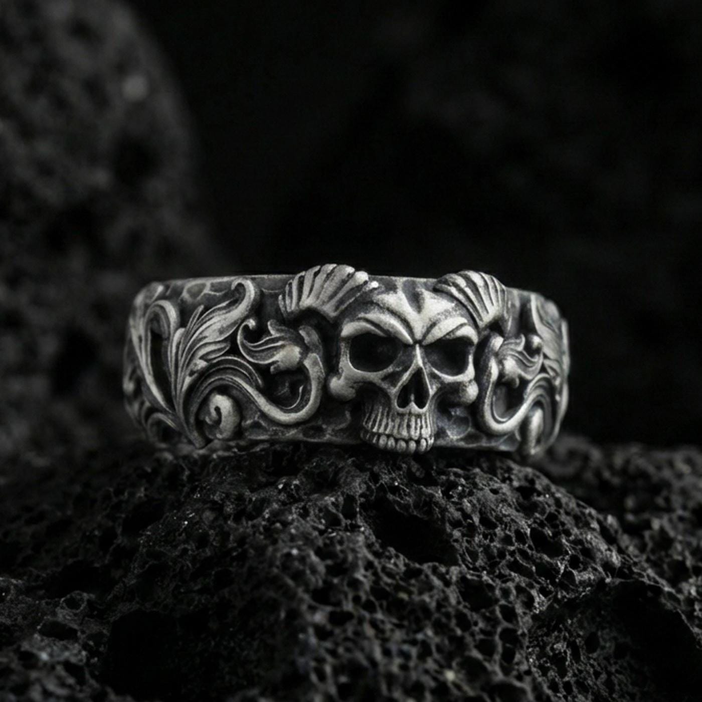 Oxidized sterling silver skull ring on volcanic stone, macro close-up highlighting horned skull relief, deep blackened recesses, and ornate scrollwork.