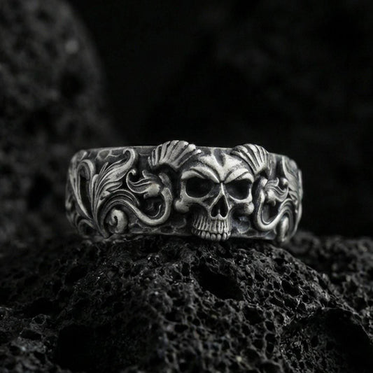 Oxidized sterling silver skull ring on volcanic stone, macro close-up highlighting horned skull relief, deep blackened recesses, and ornate scrollwork.