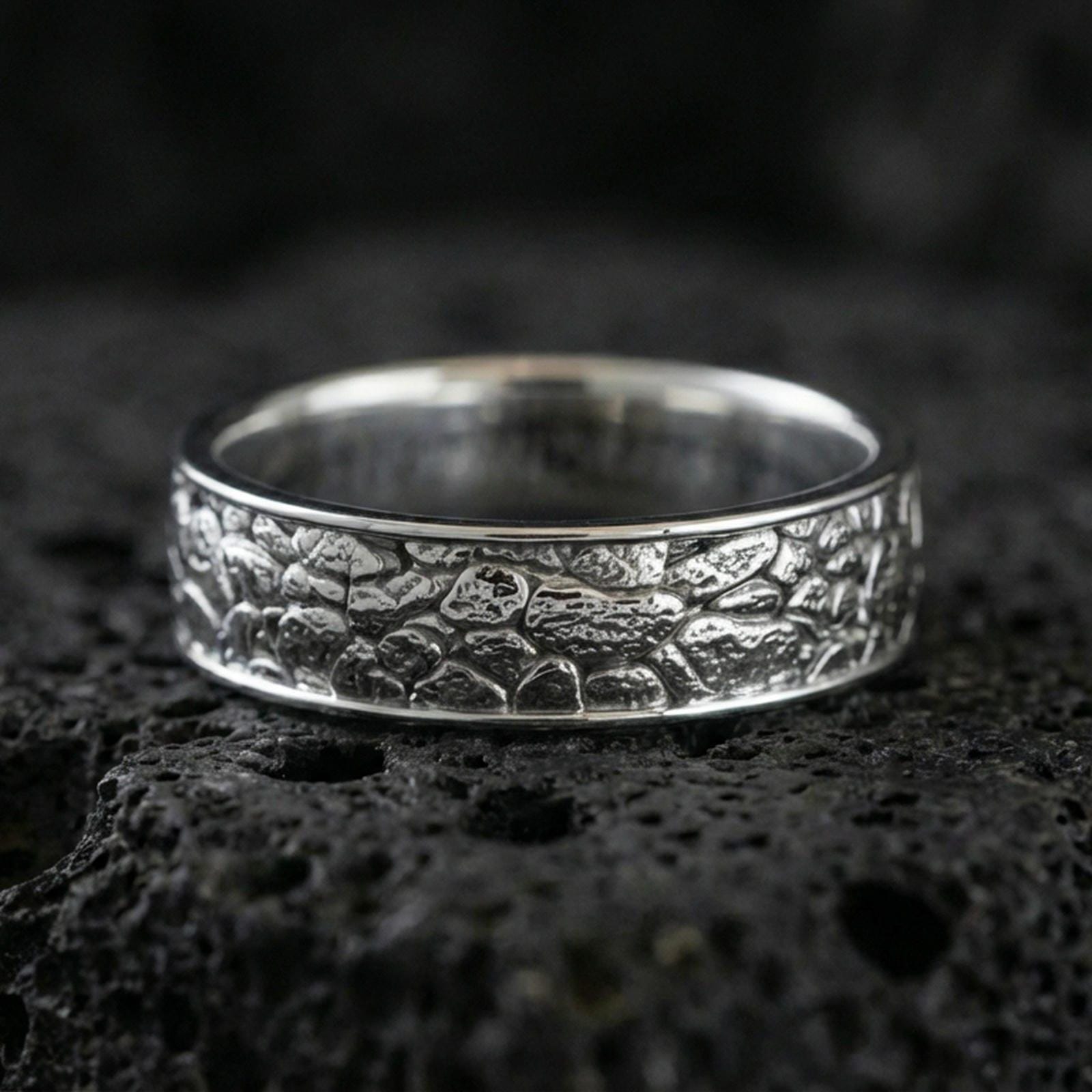 Polished silver pebble texture band ring on volcanic stone, crisp macro photo emphasizing bright shine, smooth edges, and textured surface detail.