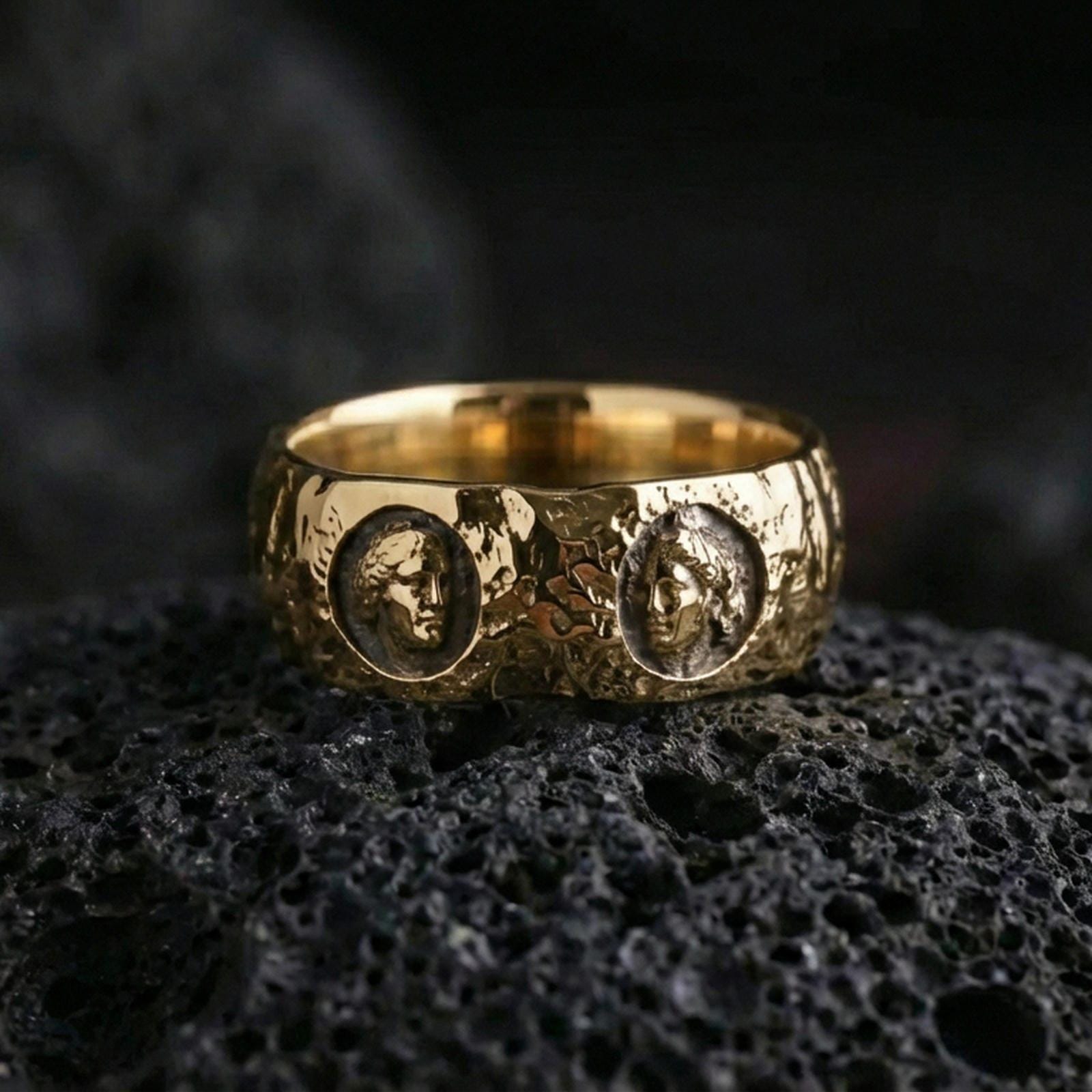 Gold plated Greek sculpture ring on volcanic stone, high-end macro photo capturing warm golden tone over the carved portraits and textured background.