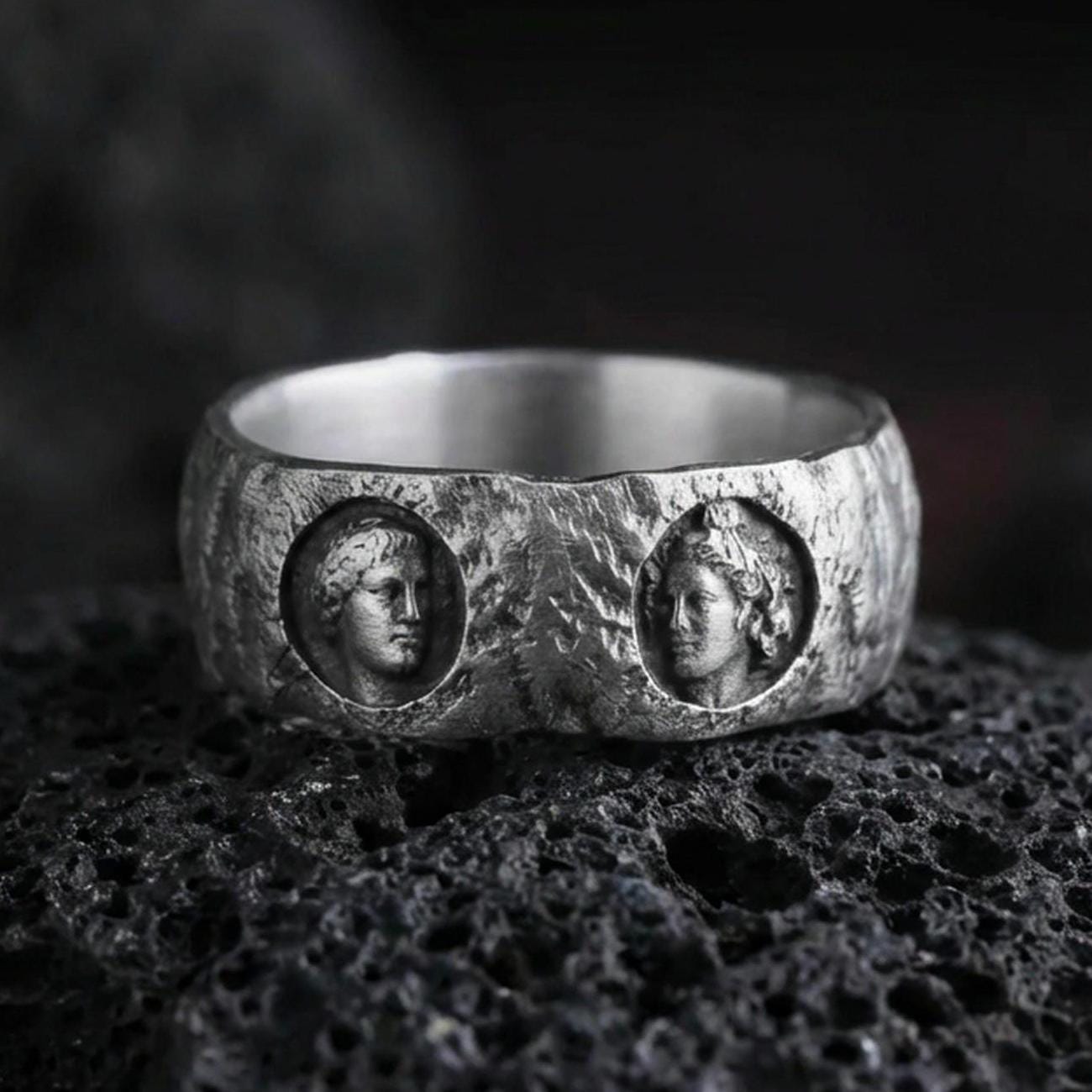Oxidized sterling silver Greek sculpture ring on volcanic stone, macro close-up focusing on the twin cameo portraits, deep shadowing, and rugged antique patina.