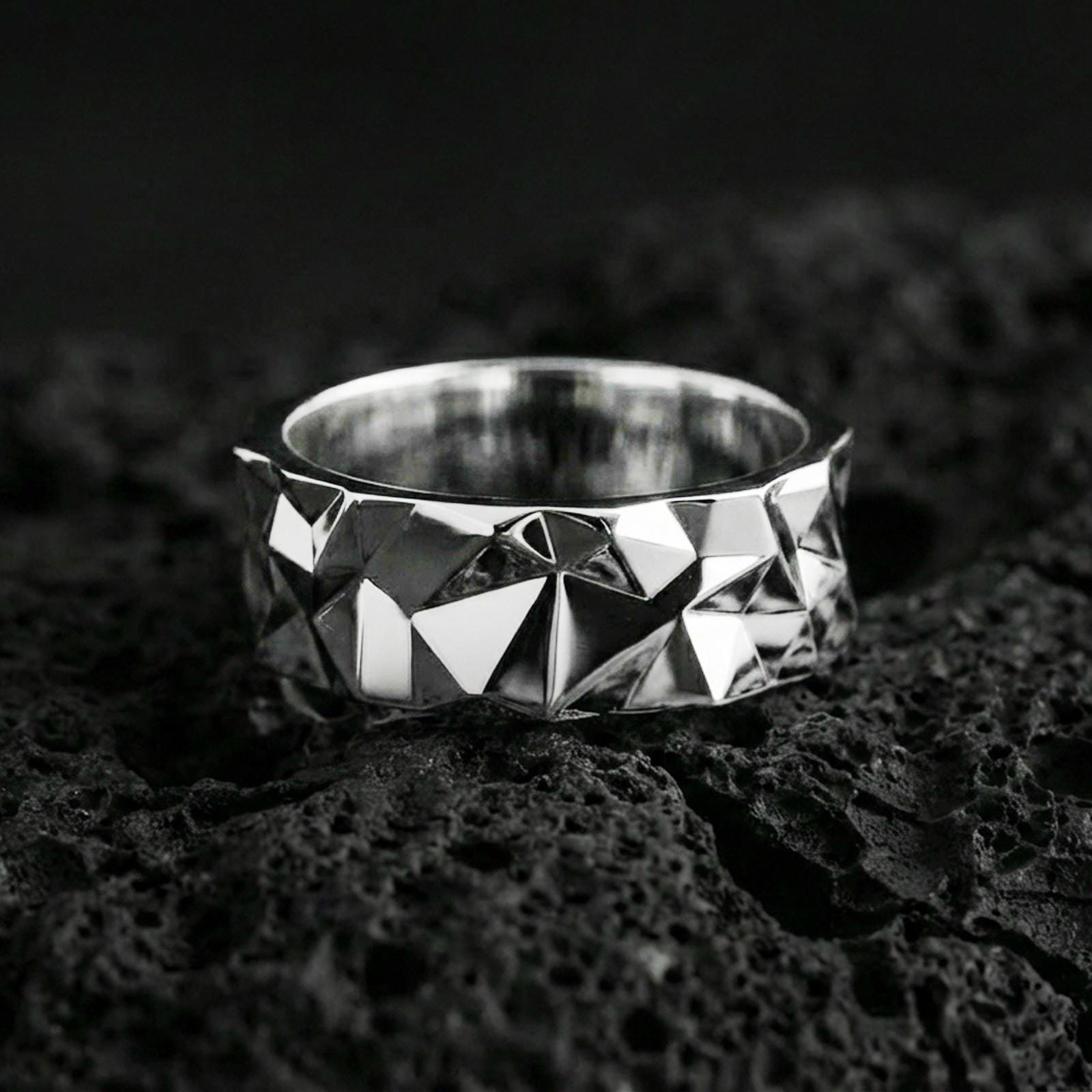 Polished silver faceted geometric band ring on volcanic stone, crisp macro shot emphasizing clean reflections, angular texture, and modern minimal form.