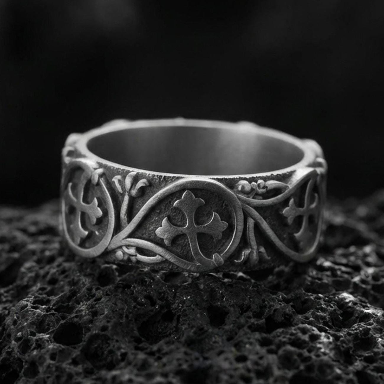 Oxidized sterling silver quatrefoil cross band ring on volcanic stone, macro close-up showing darkened recessed background and raised cross medallion details.