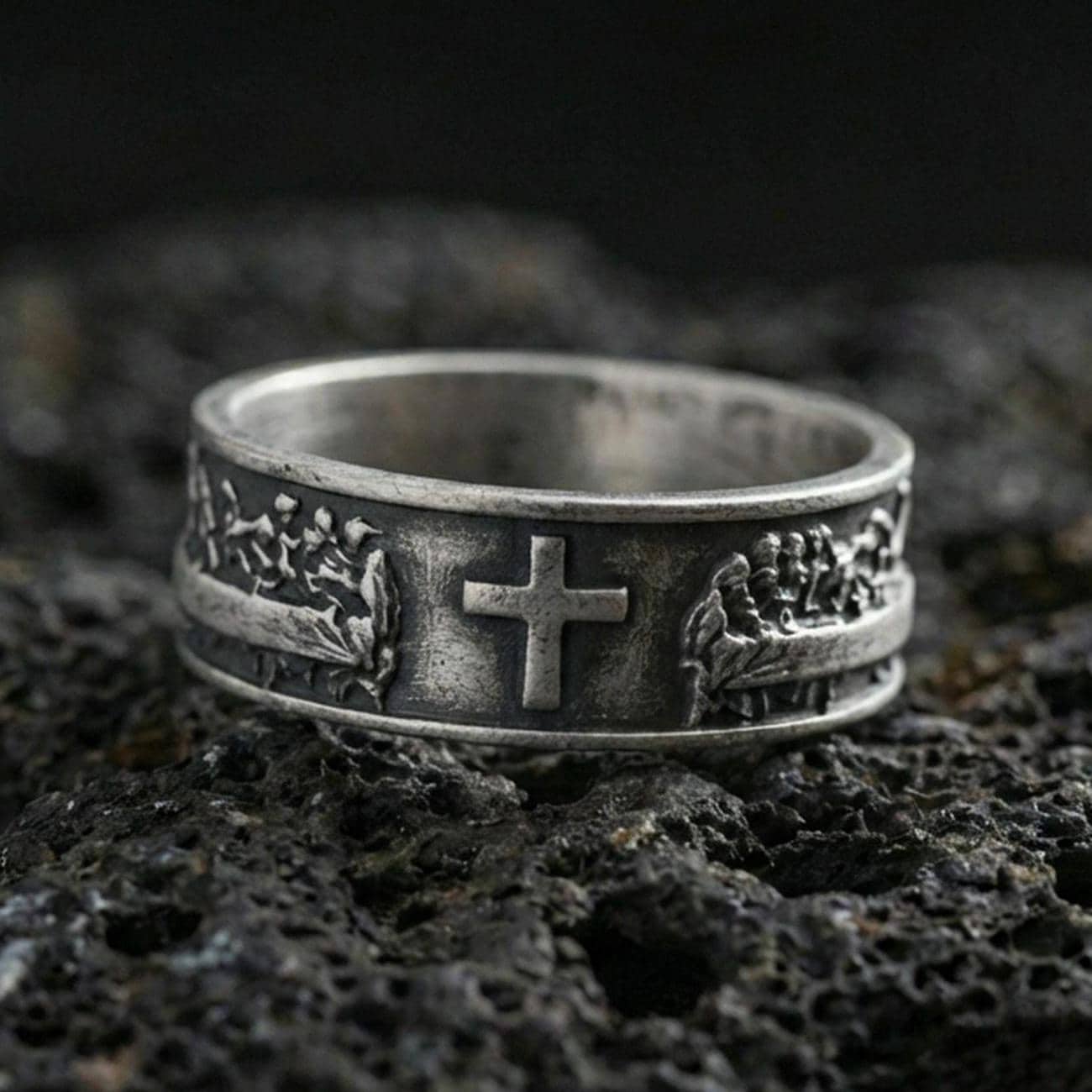 Oxidized sterling silver cross band ring on volcanic stone, macro close-up highlighting darkened recesses, raised cross centerpiece, and crisp relief texture.