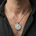 Load image into Gallery viewer, Polished silver Saint Benedict medal with engraved symbols and saint relief worn on the neck in a close lifestyle frame, highlighting the circular silhouette and sacred detailing
