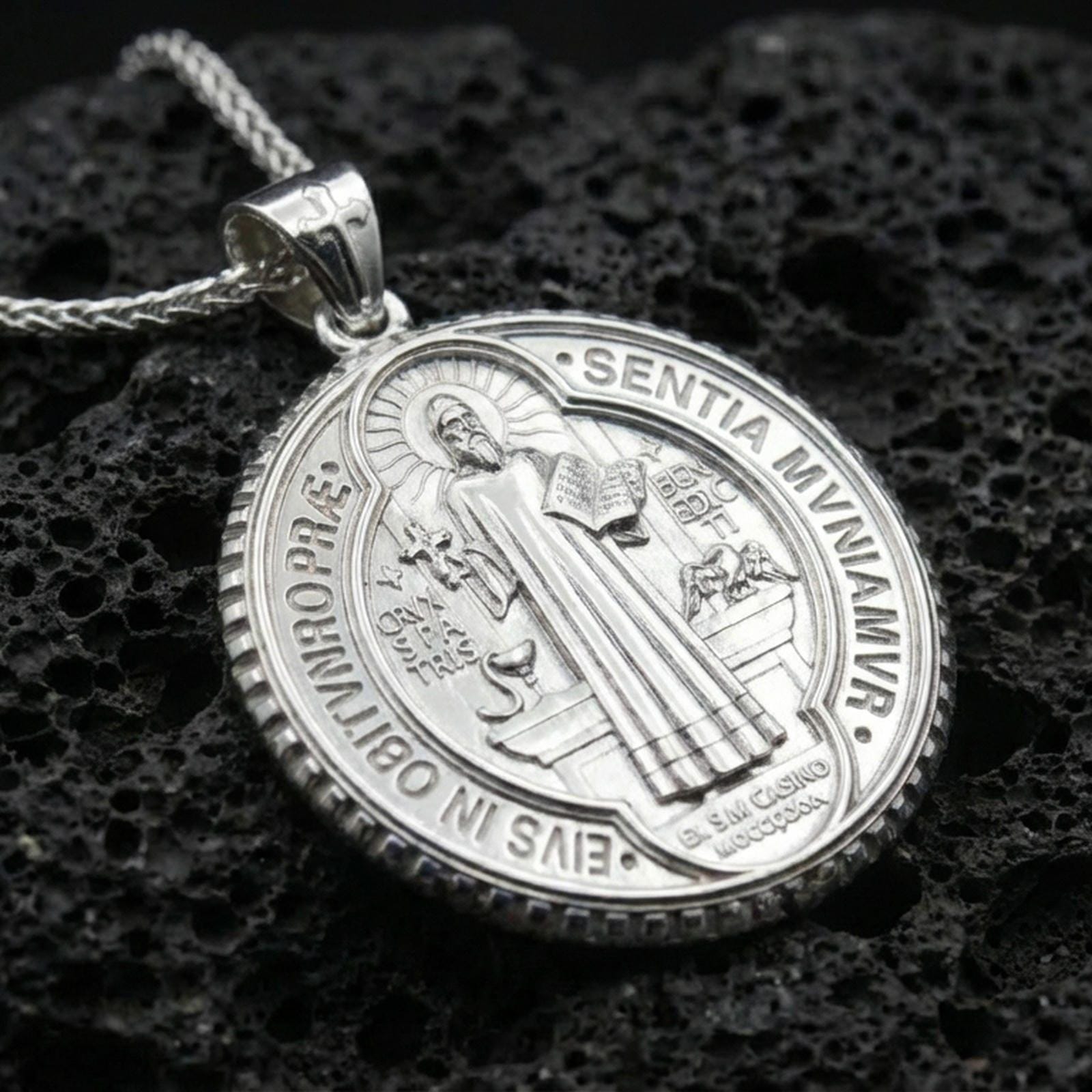 Polished silver Saint Benedict medal with detailed saint relief, engraved Benedictine symbols, and circular frame placed on textured volcanic rock in a premium studio product shot