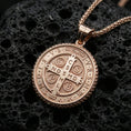 Load image into Gallery viewer, Rose gold plated Saint Benedict necklace with circular medal design, symbolic cross center, and engraved protective inscription resting on volcanic rock in an elegant close-up product presentation
