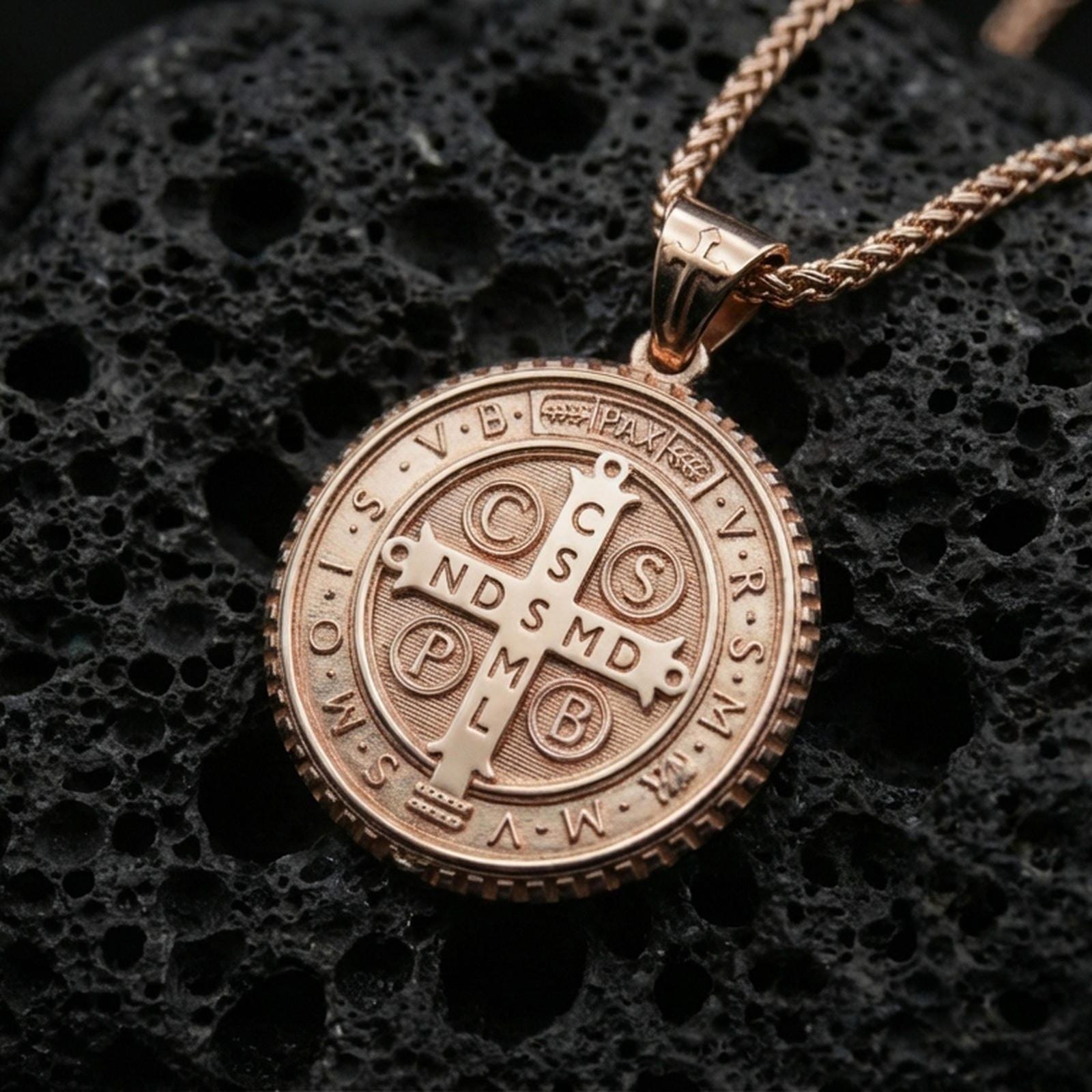 Rose gold plated Saint Benedict necklace with circular medal design, symbolic cross center, and engraved protective inscription resting on volcanic rock in an elegant close-up product presentation
