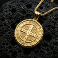 Load image into Gallery viewer, Gold plated Saint Benedict medallion with sacred cross motif, engraved devotional lettering, and classic round silhouette showcased on black volcanic rock in a refined macro jewelry image
