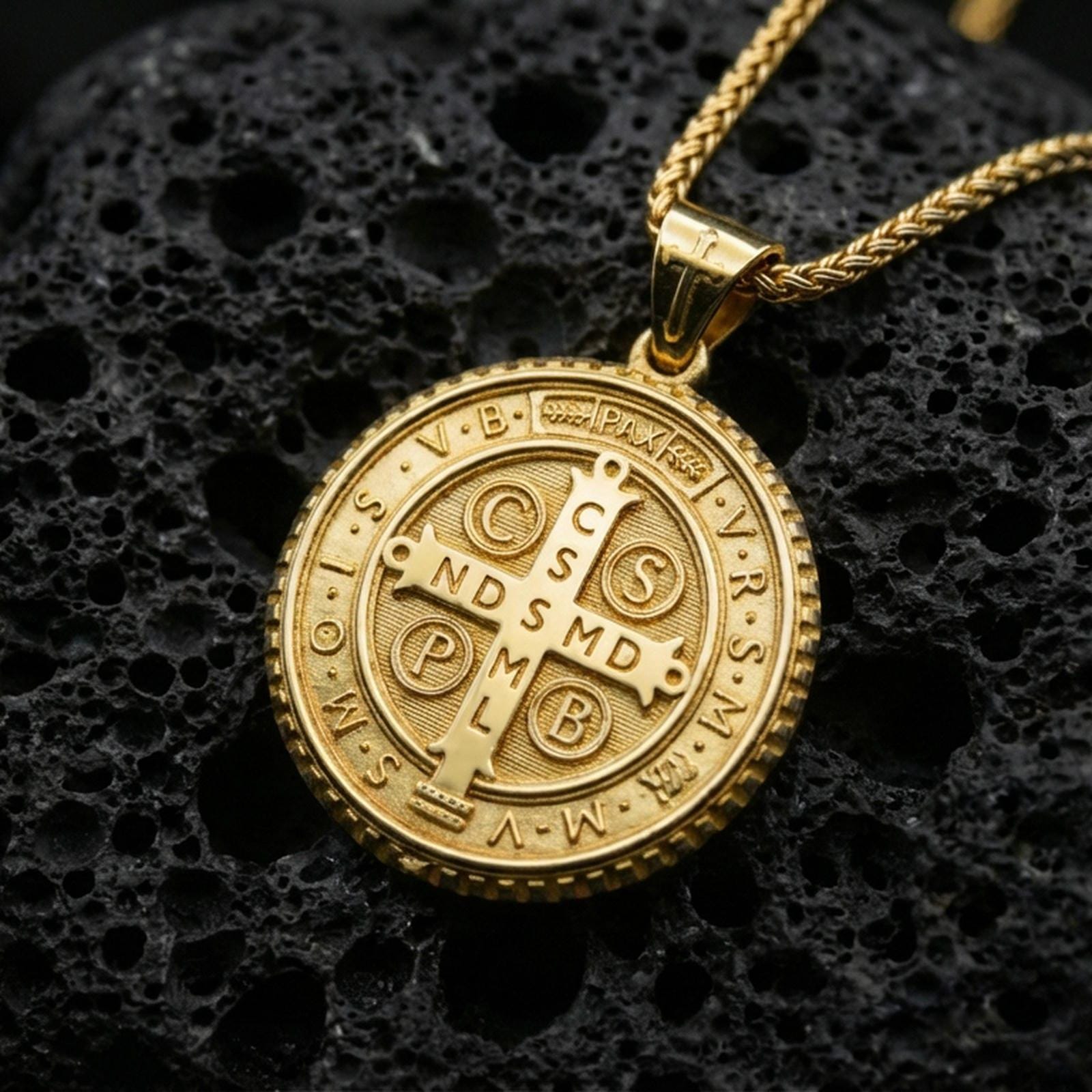 Gold plated Saint Benedict medallion with sacred cross motif, engraved devotional lettering, and classic round silhouette showcased on black volcanic rock in a refined macro jewelry image