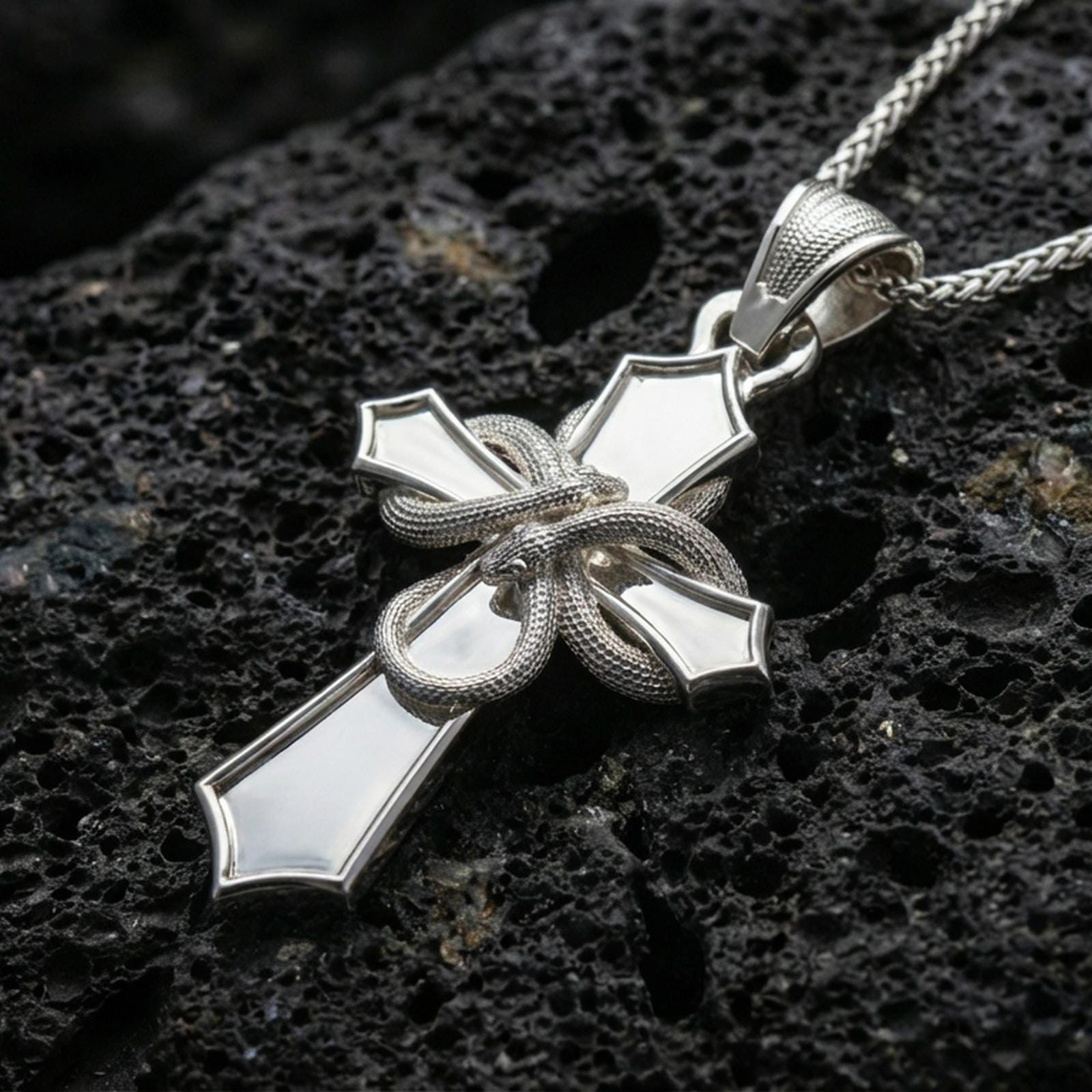 Polished silver serpent cross necklace with sculpted snake relief, flared cross ends, and dramatic symbolic design placed on textured volcanic rock in a premium studio product shot