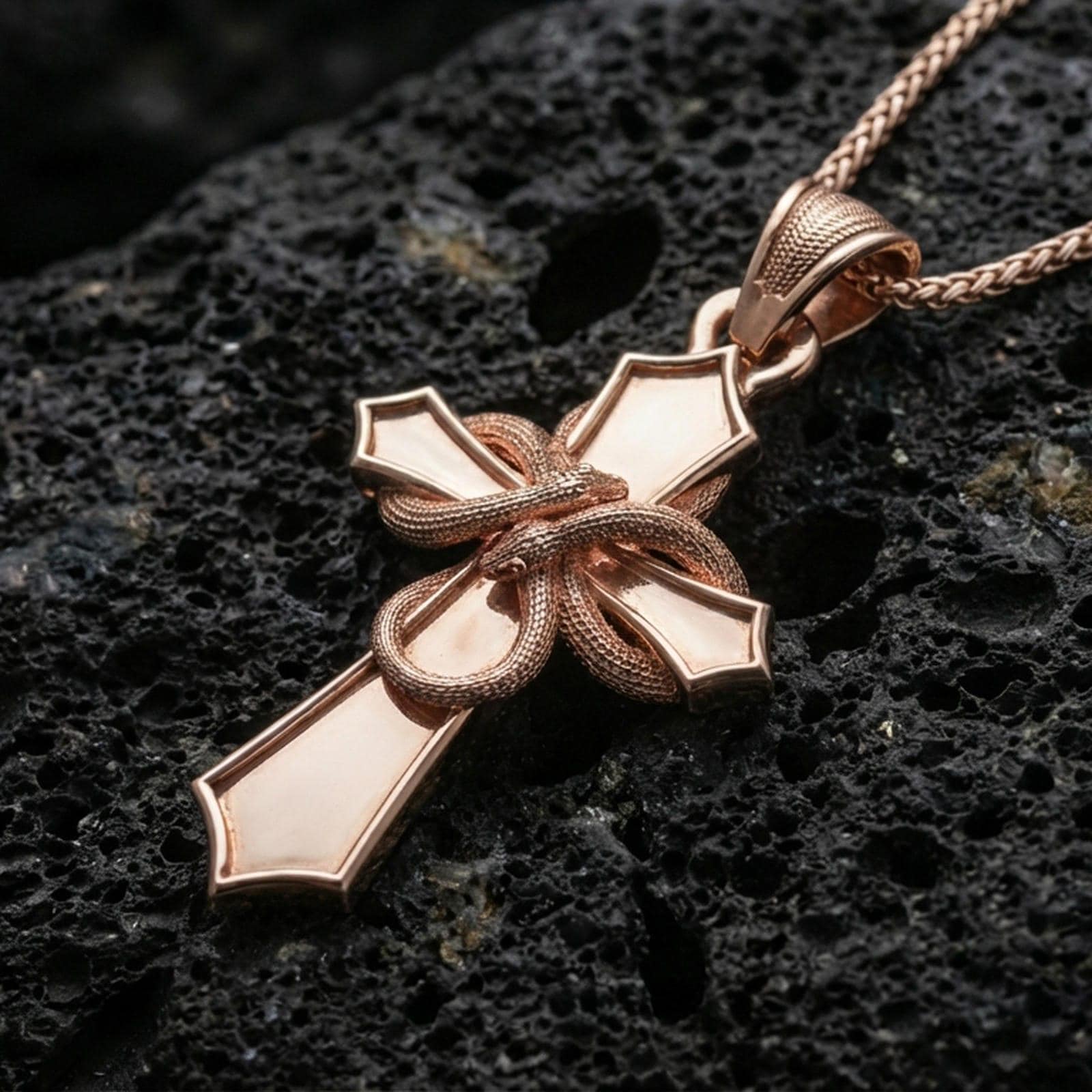 Rose gold plated gothic cross pendant with wrapped serpent motif, polished cross silhouette, and striking sculptural detail resting on volcanic rock in an elegant close-up product presentation