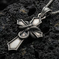 Load image into Gallery viewer, Oxidized cross pendant with wrapped snake detail and bold gothic silhouette displayed on dark volcanic rock in a close-up luxury jewelry composition
