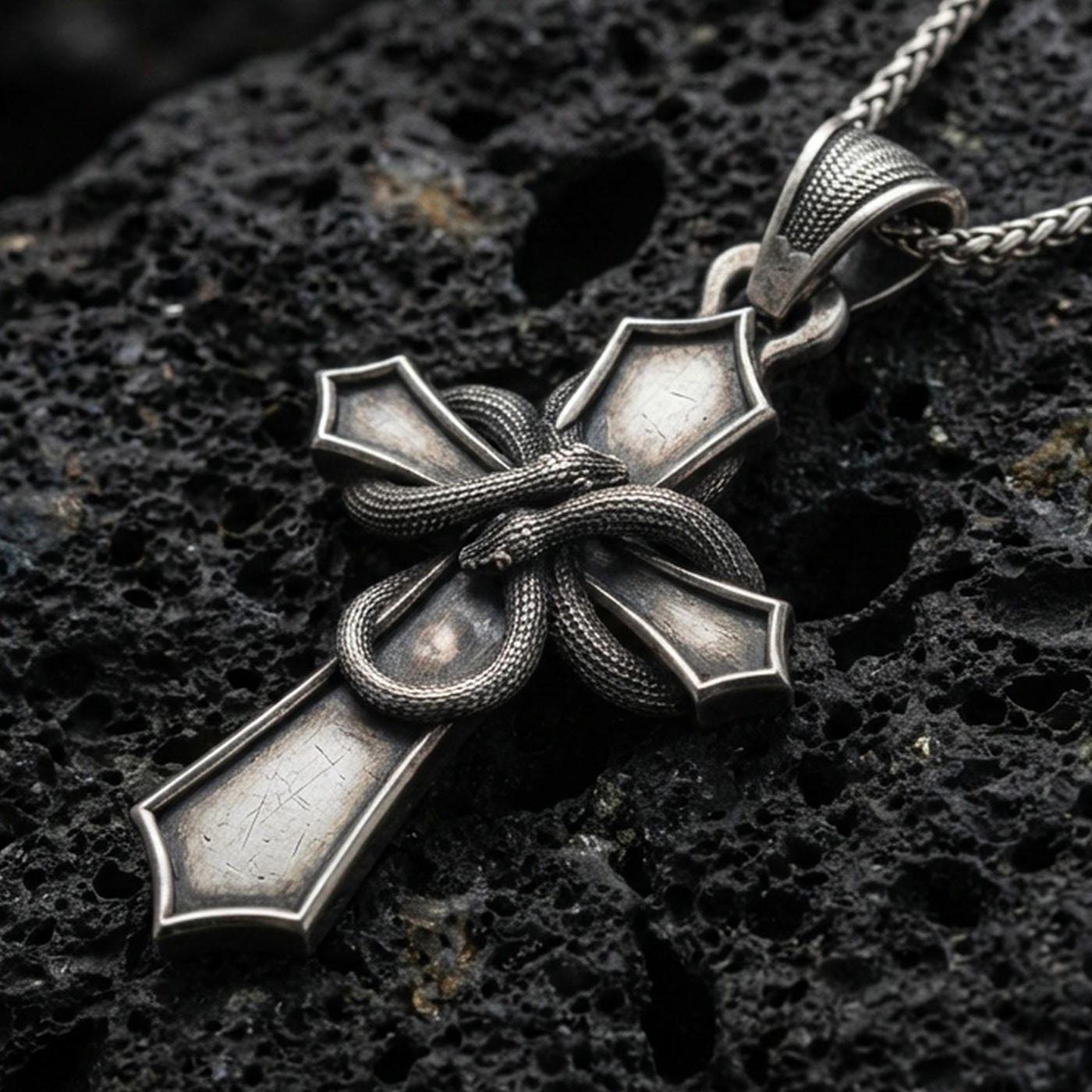 Oxidized cross pendant with wrapped snake detail and bold gothic silhouette displayed on dark volcanic rock in a close-up luxury jewelry composition
