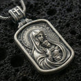 Load image into Gallery viewer, Oxidized Virgin Mary and Baby Jesus pendant with halo details, framed devotional portrait, and rounded rectangular icon design displayed on dark volcanic rock in a close-up luxury jewelry composition

