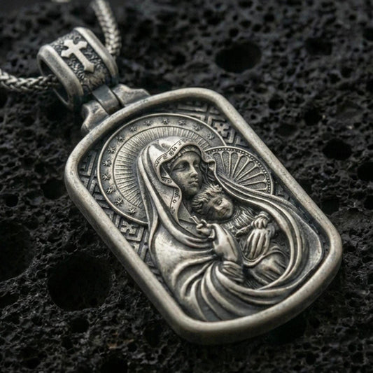 Oxidized Virgin Mary and Baby Jesus pendant with halo details, framed devotional portrait, and rounded rectangular icon design displayed on dark volcanic rock in a close-up luxury jewelry composition