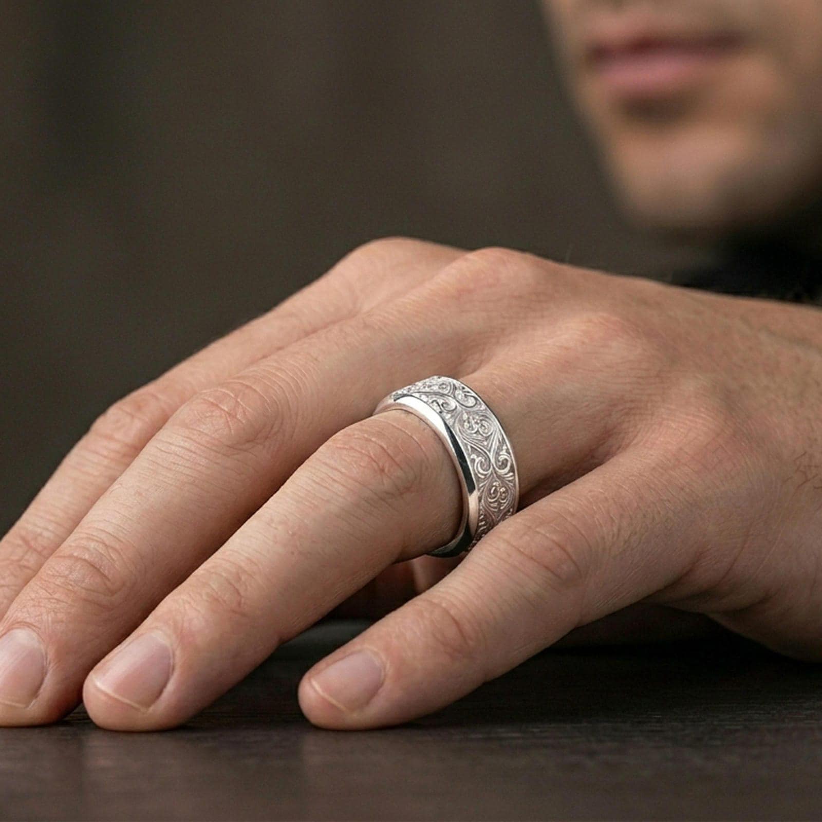 Ornate scrollwork sterling silver band ring worn on a finger, polished silver look with smooth reflections highlighting the engraved floral-scroll texture.