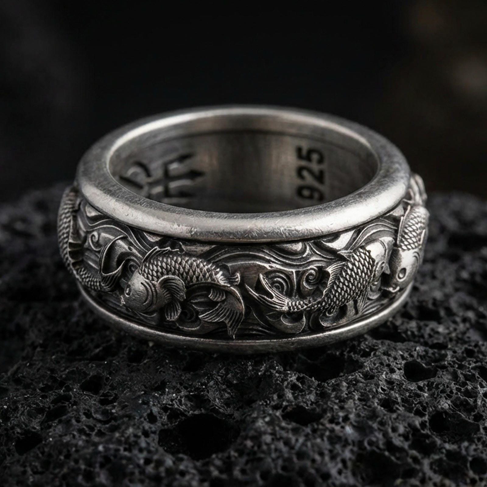 Koi fish sterling silver band ring on black volcanic stone, photographed with an oxidized finish to emphasize contrast in the carved scales and flowing water waves.