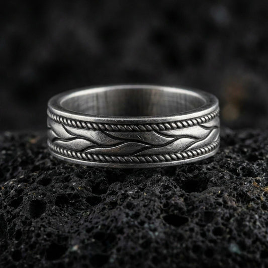 Sterling silver wave band ring on black volcanic stone, photographed with an oxidized patina that deepens the recessed swirl details and rope-edge borders.