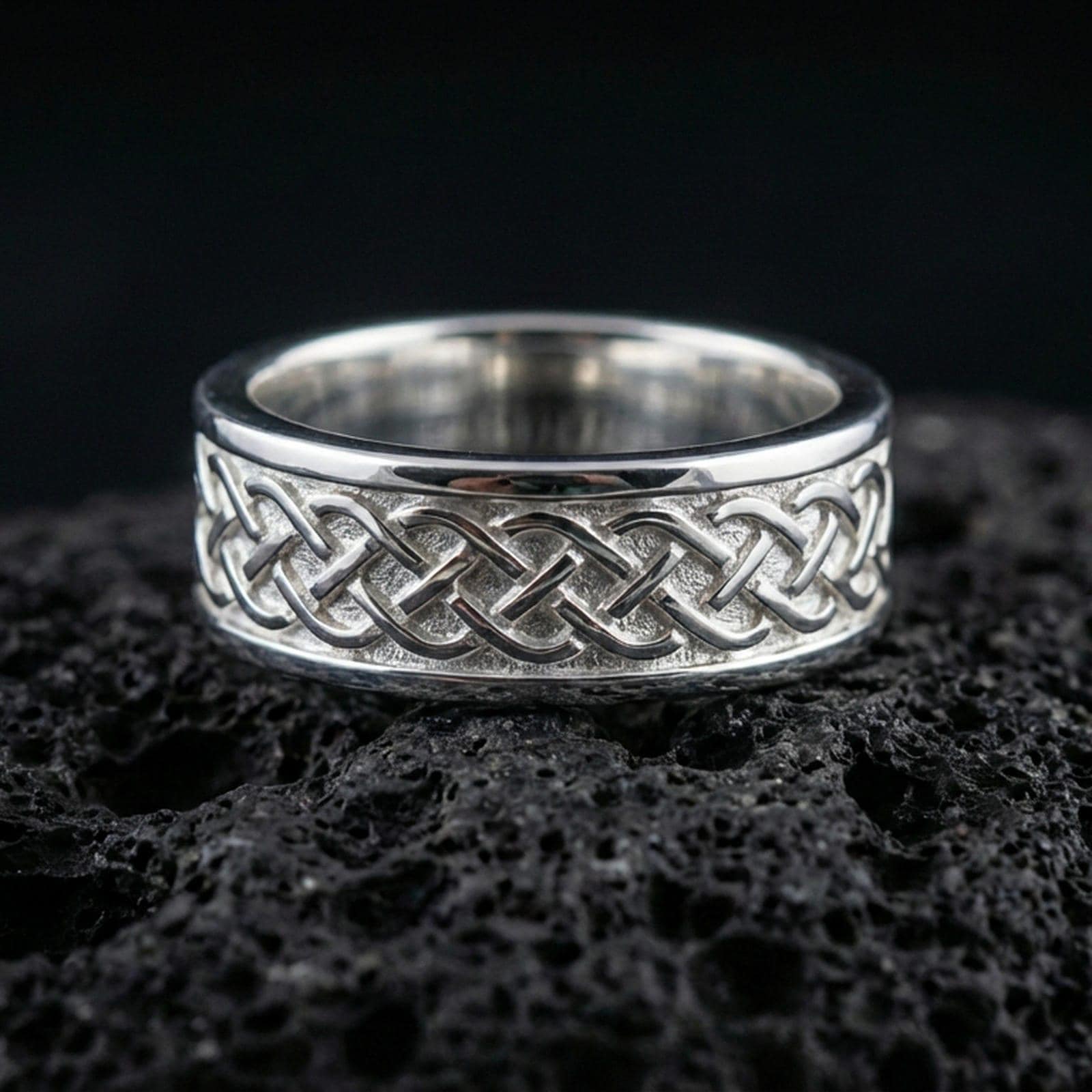 Sterling silver Celtic knot band ring on black volcanic stone, shown in a polished silver look with clean highlights across the interlaced knot pattern and smooth edges.