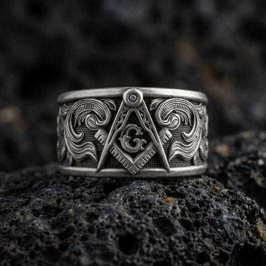 Sterling silver Masonic square and compass band ring on black volcanic stone, photographed with an oxidized patina that darkens the recessed background and sharpens the raised emblem details.