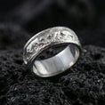 Load image into Gallery viewer, Sterling silver band ring photographed on black volcanic stone, shown in a polished silver finish with crisp highlights across the curved edges and sculpted figures.

